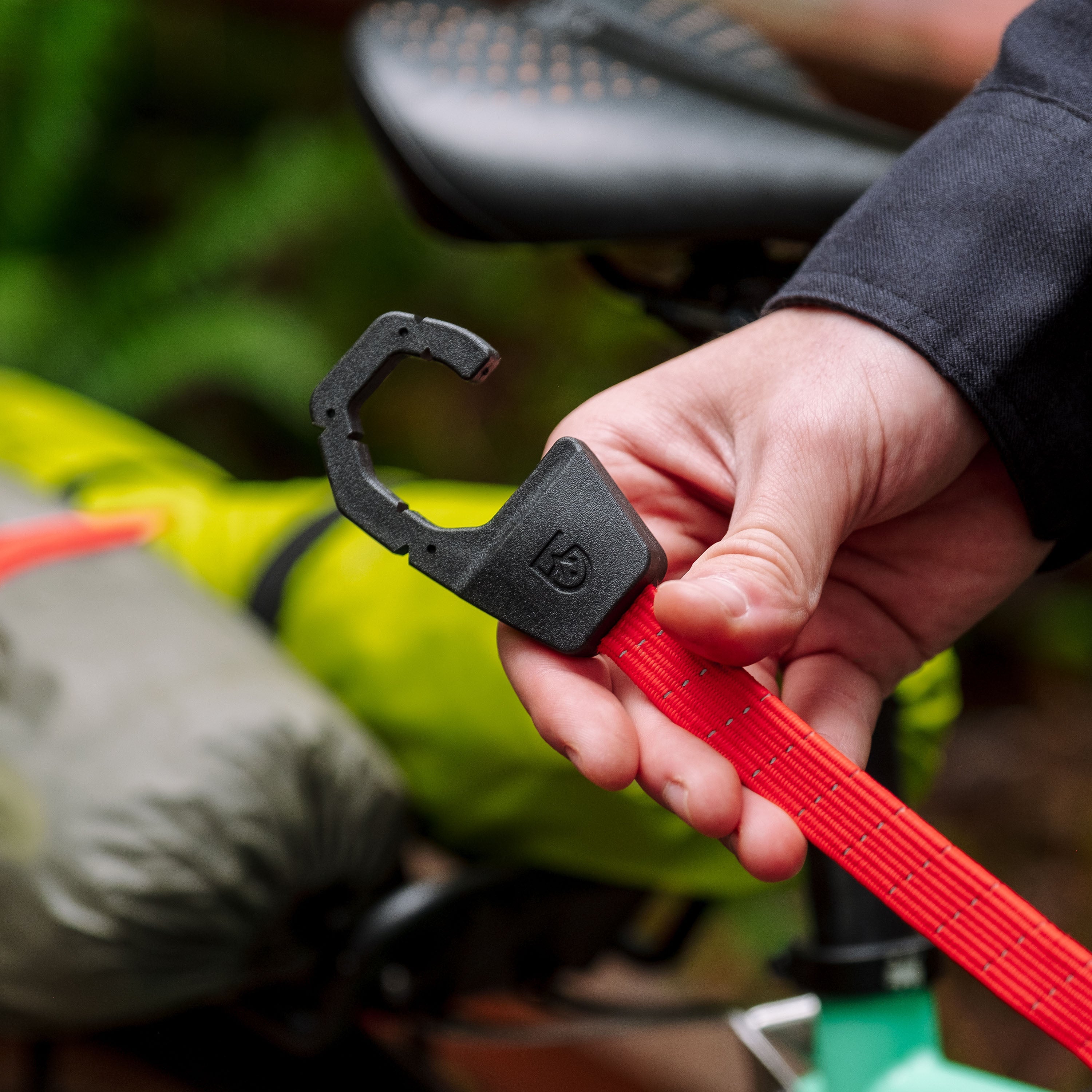 Close up of a person holding a red Holdfast Strap bungee with an over-molded steel hook with a blurry bike and bike bags in the background