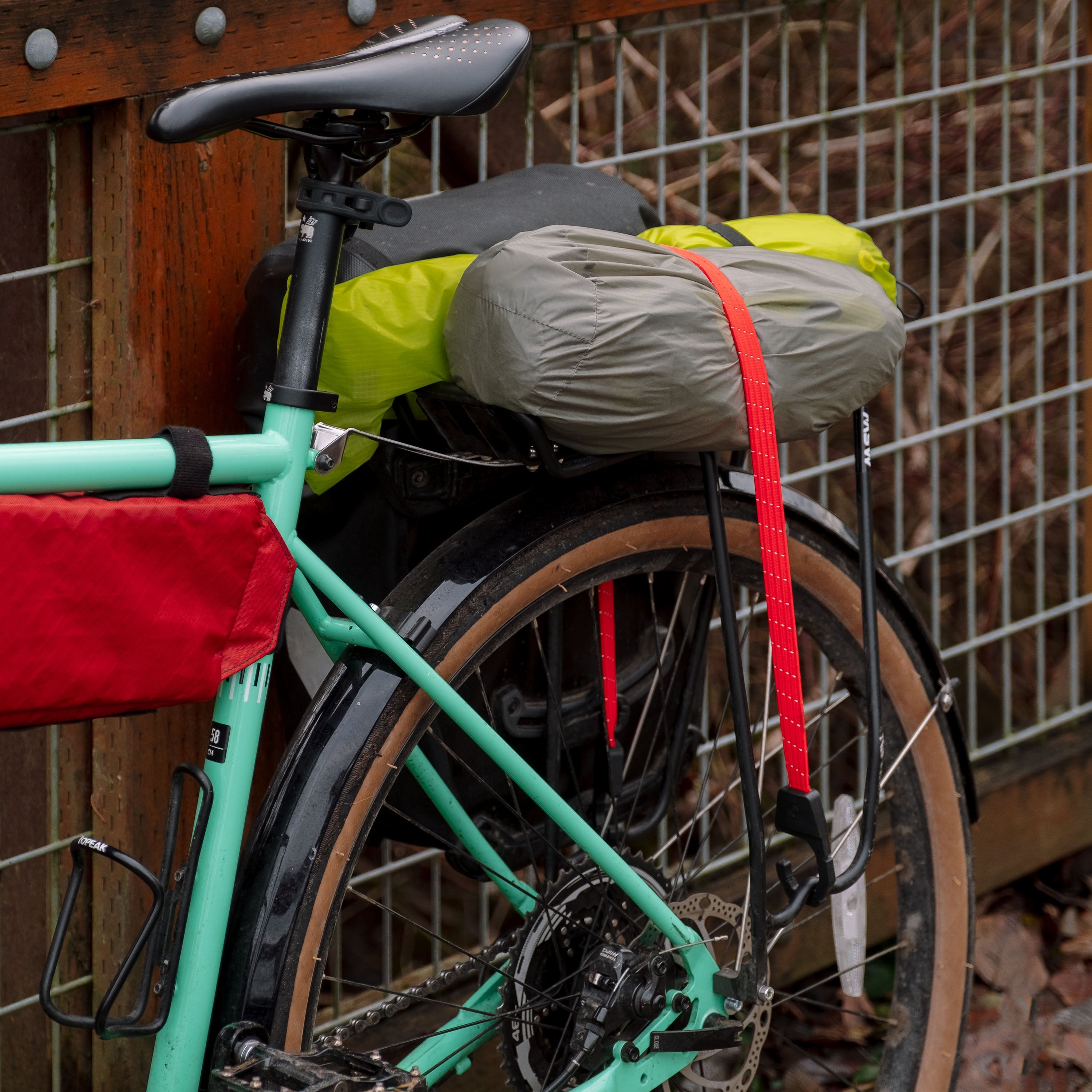 Holdfast strap holding a tent and tent rainfly onto a rack on a teal gravel bike, resting against a wooden and metal fence with brown foliage in the background